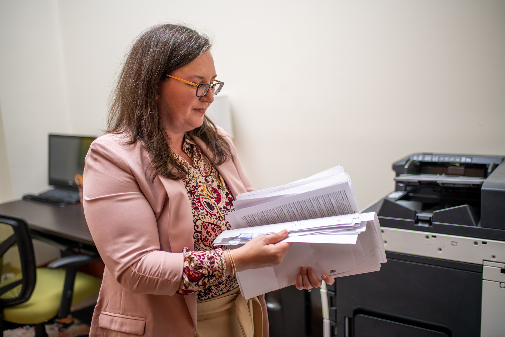 A middle-aged white woman with glasses, wearing a business jacket, holds a large stack of papers. 