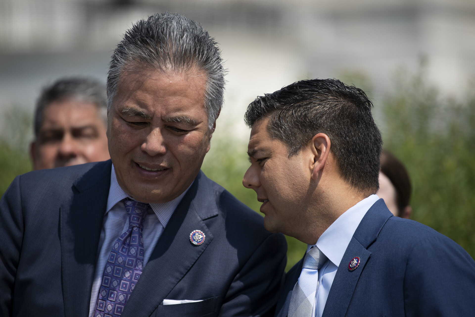 A close-up of two middle-aged men in blue suits outside on a sunny day, both with trim, dark haircuts. The man on the right, who appears Latino, speaks into the ear of the other, who appears Asian. 