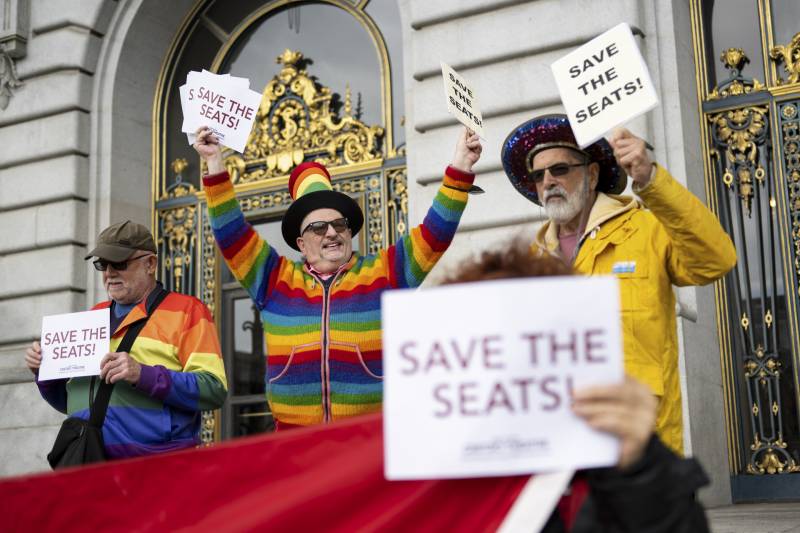 Several people in rainbow patterned clothing hold signs that read "save the seats" in front of ornate gold-leafed doors.