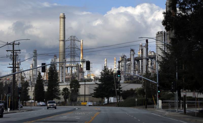 The silver smoke stacks of a large oil refinery loom above a city street where vehicles and a stoplight appear in the foreground