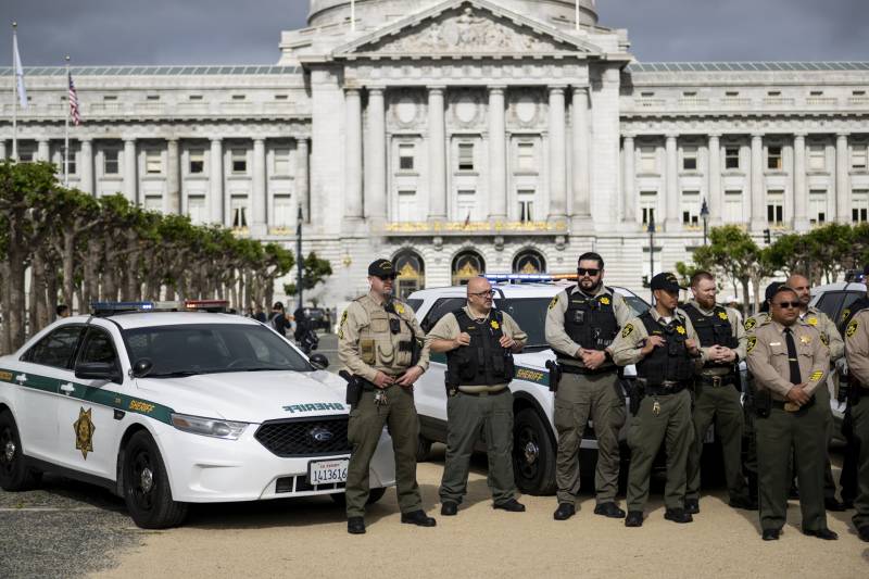 A group of half a dozen law enforcement officers stand in front of squad cars parked in an open area in front of an ornate columned building.