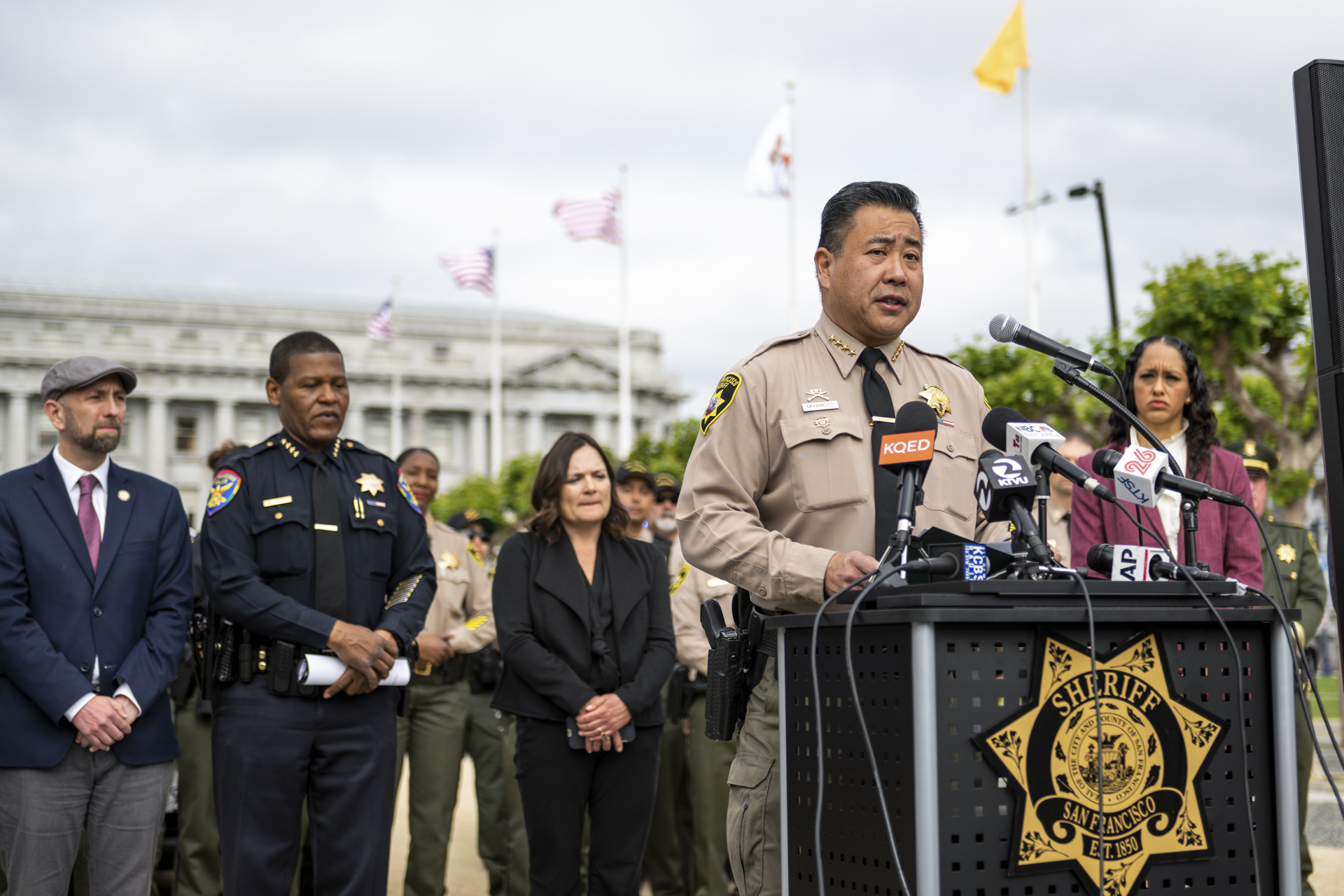 A man in an official uniform with a starred badge pinned to it speaks into an array of microphones from an outdoor lectern, flanked by law enforcement officers and others.