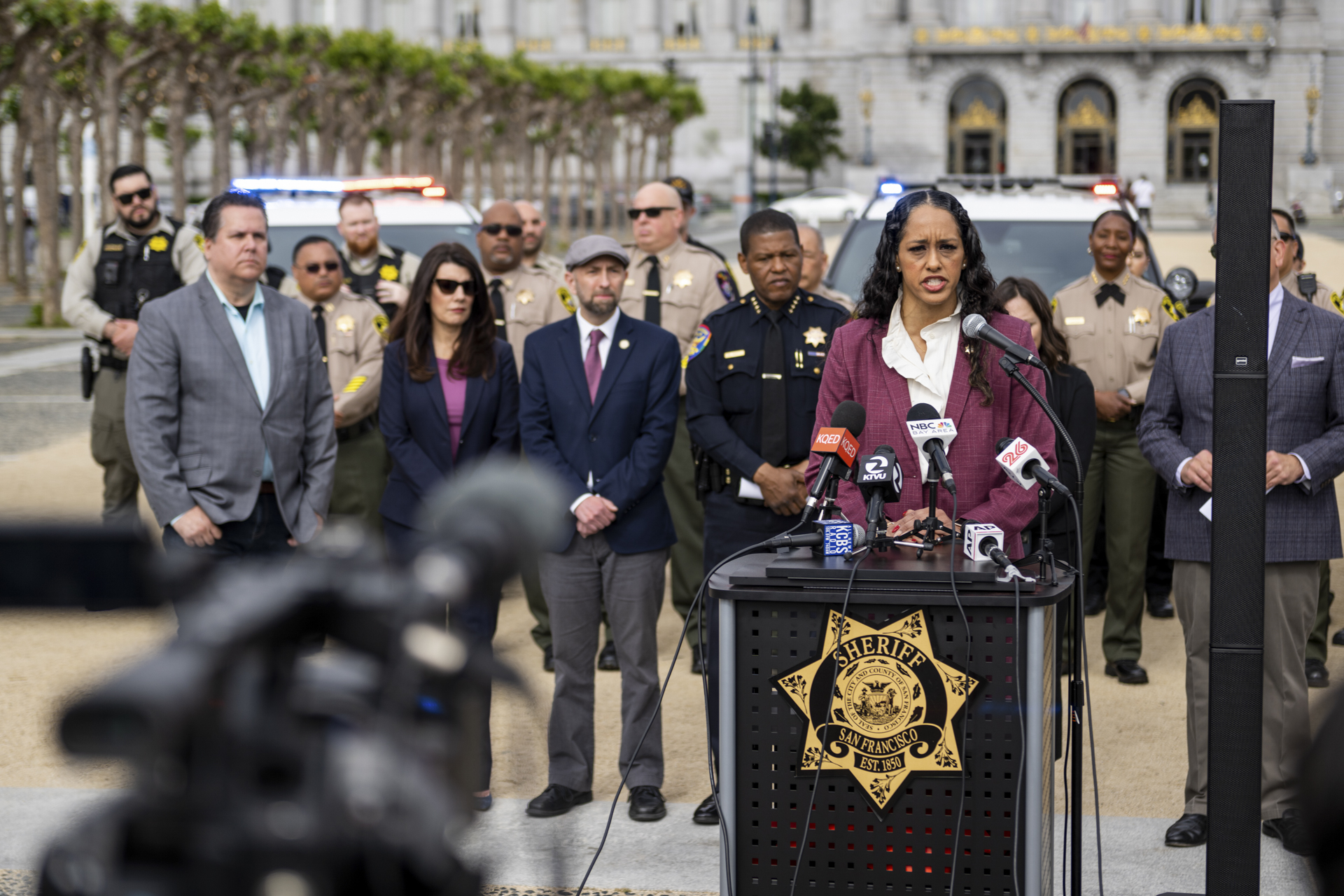 A woman in a purple suit speaks into an array of microphones from an outdoor lectern, flanked by law enforcement officers and others.