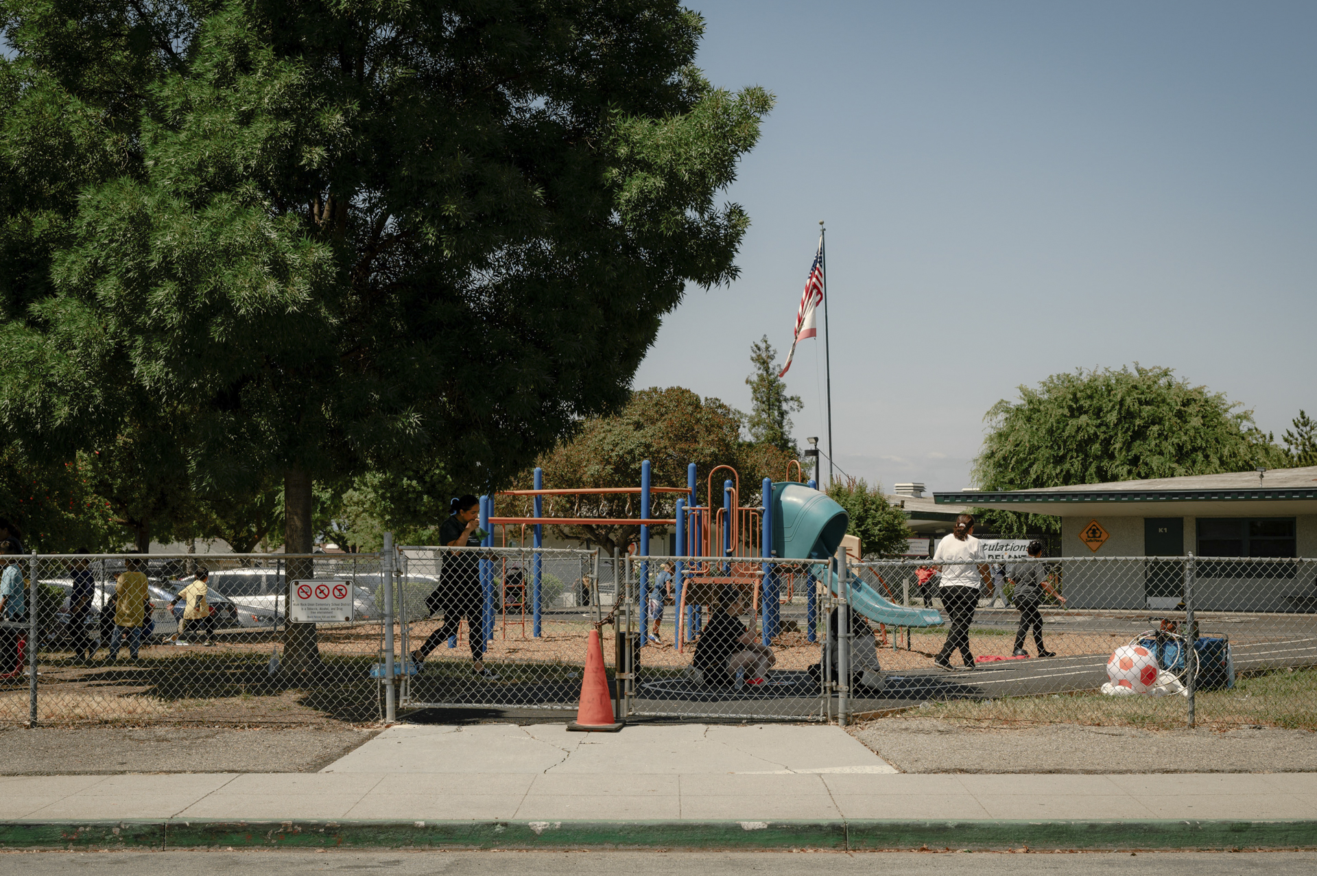 A children and adults circulate beside a playground behind a chainlink fence.