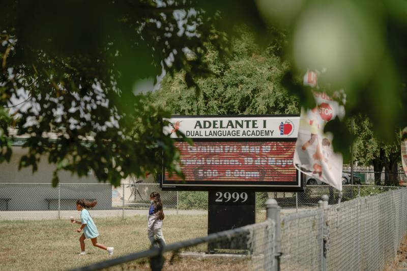 Children play on grass behind a chainlink- fence beside a sign reading "Adelante I: Dual Language Academy."