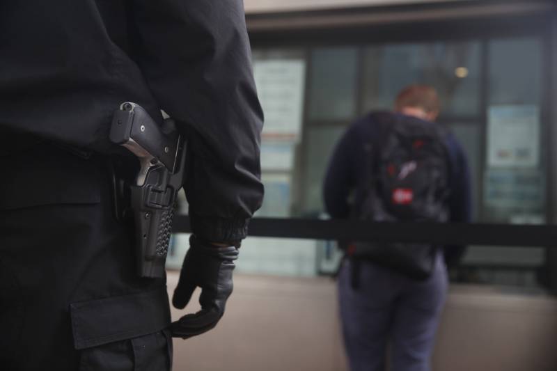 a black-clad security guard's outstretched arm and leg are seen from behind in the left foreground, with a gun clearly visible on their hip, as a pedestrian stands with their back turned to the camera in the background