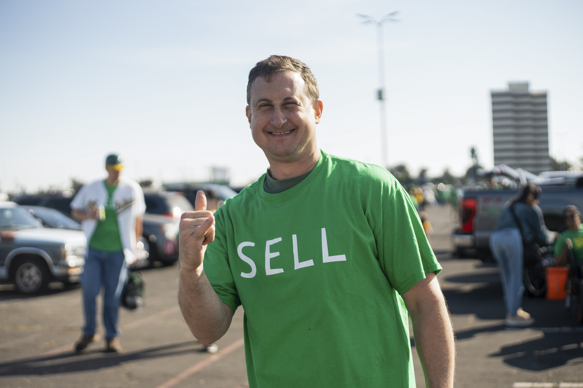 A man in a lime green shirt that reads "sell" points and smiles at the camera while standing in a parking lot.