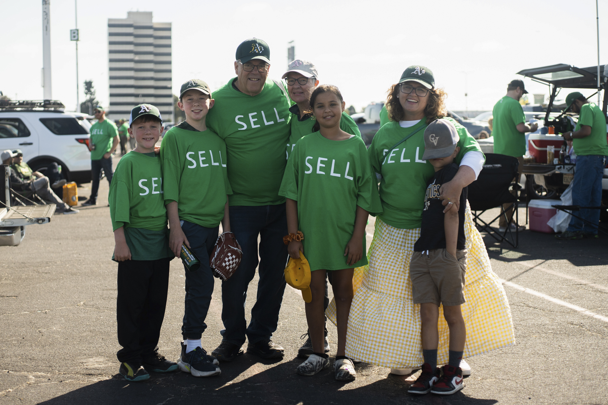 A group of seven people stand together to take a photo in an outdoor parking lot, all but one wear t-shirts reading "sell" and several wear Oakland A's hats.