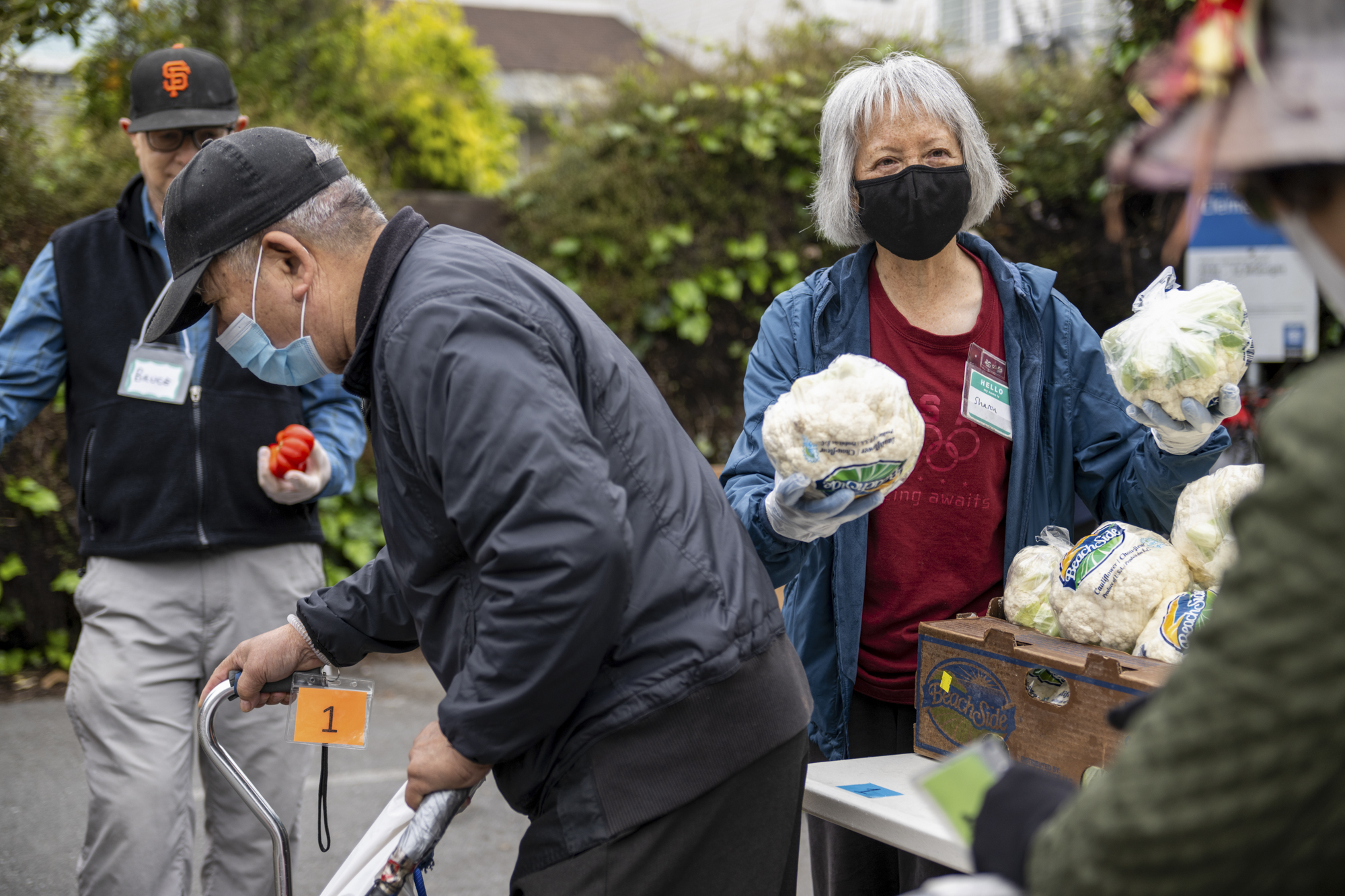 A woman with gray hair wearing a black face mask holds out cauliflower heads as a man using a can walks past in a paved outdoor area.