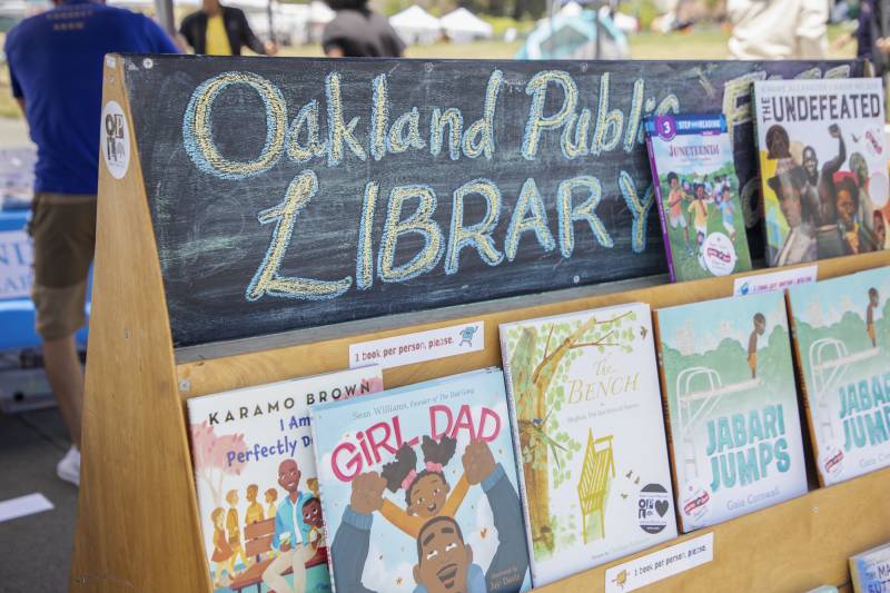 A wooden bookshelf with three rows of children's books, covers facing out, and a chalk sign at the top saying, "Oakland Public Library."