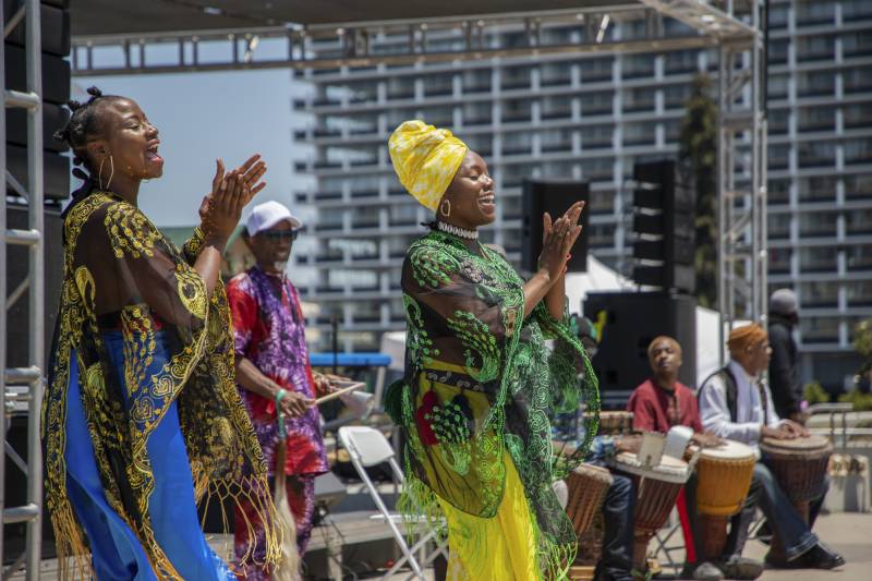 Two Black women, one in Bantu knots and another in a bright yellow head wrap, both in long, flowing, colorful clothing, dance alongside a drum circle on an outdoor stage.