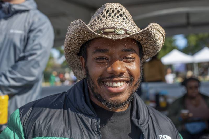 A Black man with a wide grin and salt and pepper in his beard and mustache smiles into the sun beneath a woven cowboy hat.