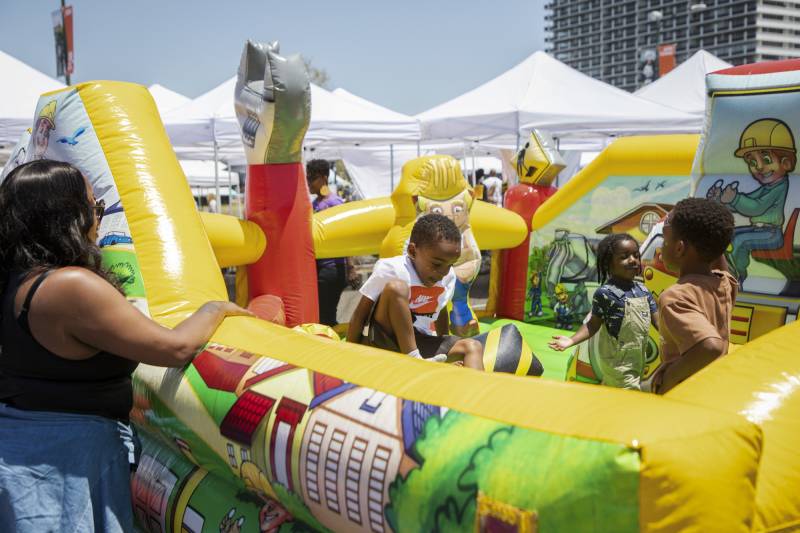 Three Black children tumble and smile in an open jumpy house that is bright yellow, with neighborhood scenes on it, as a Black woman with long black hair and sunglasses rests her right arm on the side and watches them.