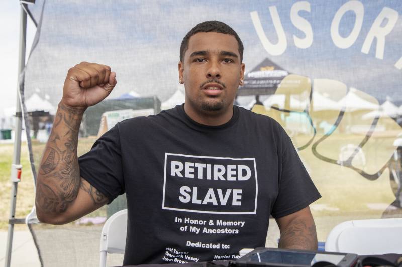 A young Black man with trim hair and goatee and tattoo sleeves, wearing a black T-shirt that says "Retired Slave / In Honor & Memory and My Ancestors," raises his right fist and smiles slightly as he looks at the camera.