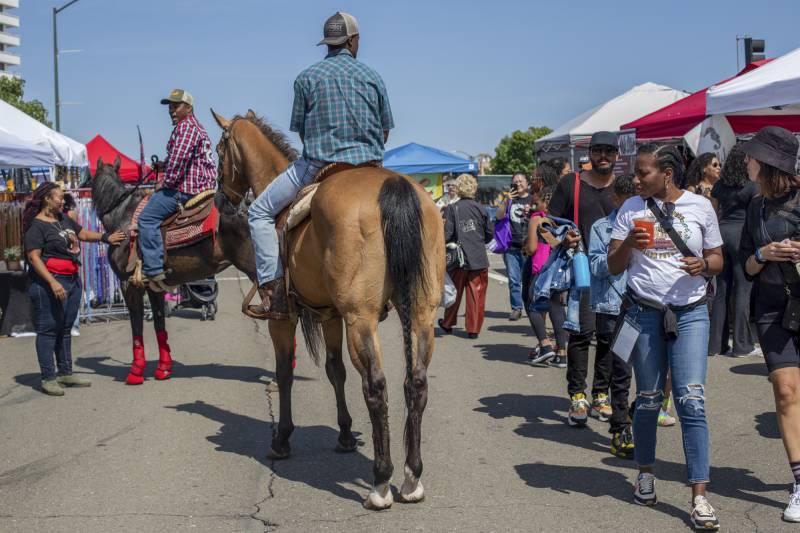 Amid white and red vendor tents, two Black men in checkered shirts, jeans, and baseball caps sit atop horses on the asphalt, both stopped to talk to people below them dressed casually and carrying drinks.