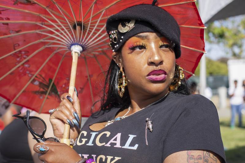 A young Black women with elaborate makeup -- pink and orange eye shadow, lined lips, '80s-style gold bamboo hoops, and a nose ring -- with long reflective blue-gray nails, holding a red parasol and wearing a black beanie with brooches on it, looks beyond the camera.