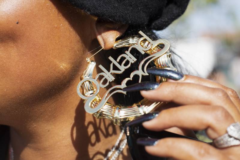 A close-up of a Black woman holding up her '80s-style bamboo earring, with the word "Oakland" across it. Her hand has long, dark gray-painted nails.