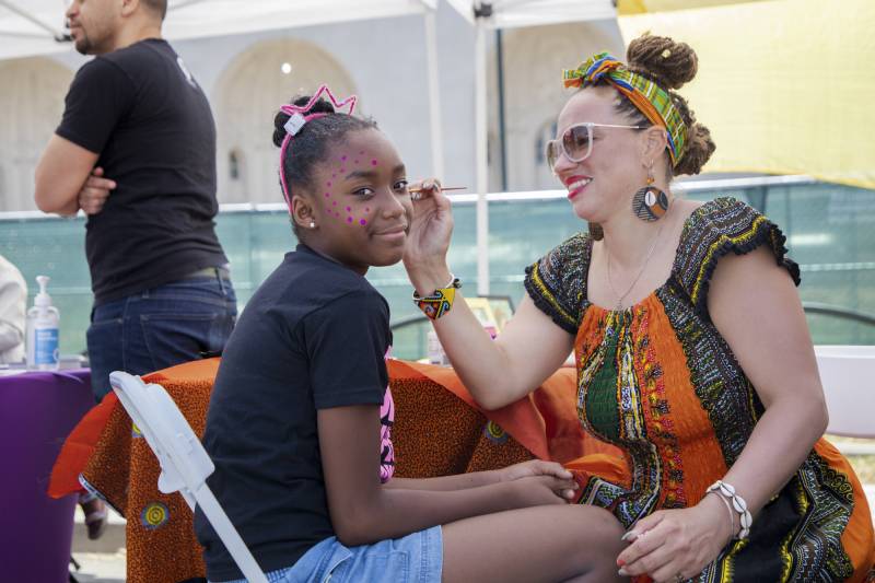 A young Black girl turns her face, smiling, to the camera, red dots decorating the right side of her face, as a Black woman in an orange and green dress and sunglasses paints the left side of her face. They sit knee to knee on folding chairs.