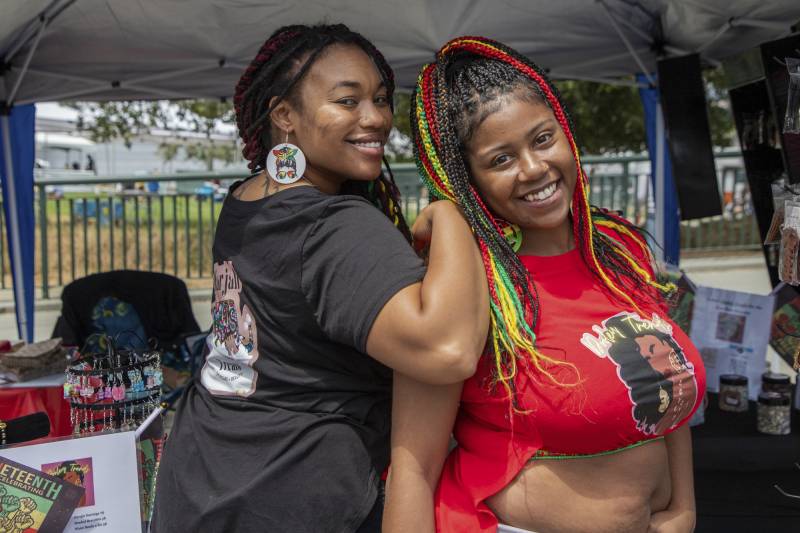Two Black woman pose, smiling at the camera. The woman on the left looks over her right shoulder, showing the graphic on the back of her T-shirt and holding her friend's shoulder with her right arm. Both have long braids; the woman on the right has red, green, and yellow braids, and wears a red crop top, also a graphic T with an image of a Black woman.