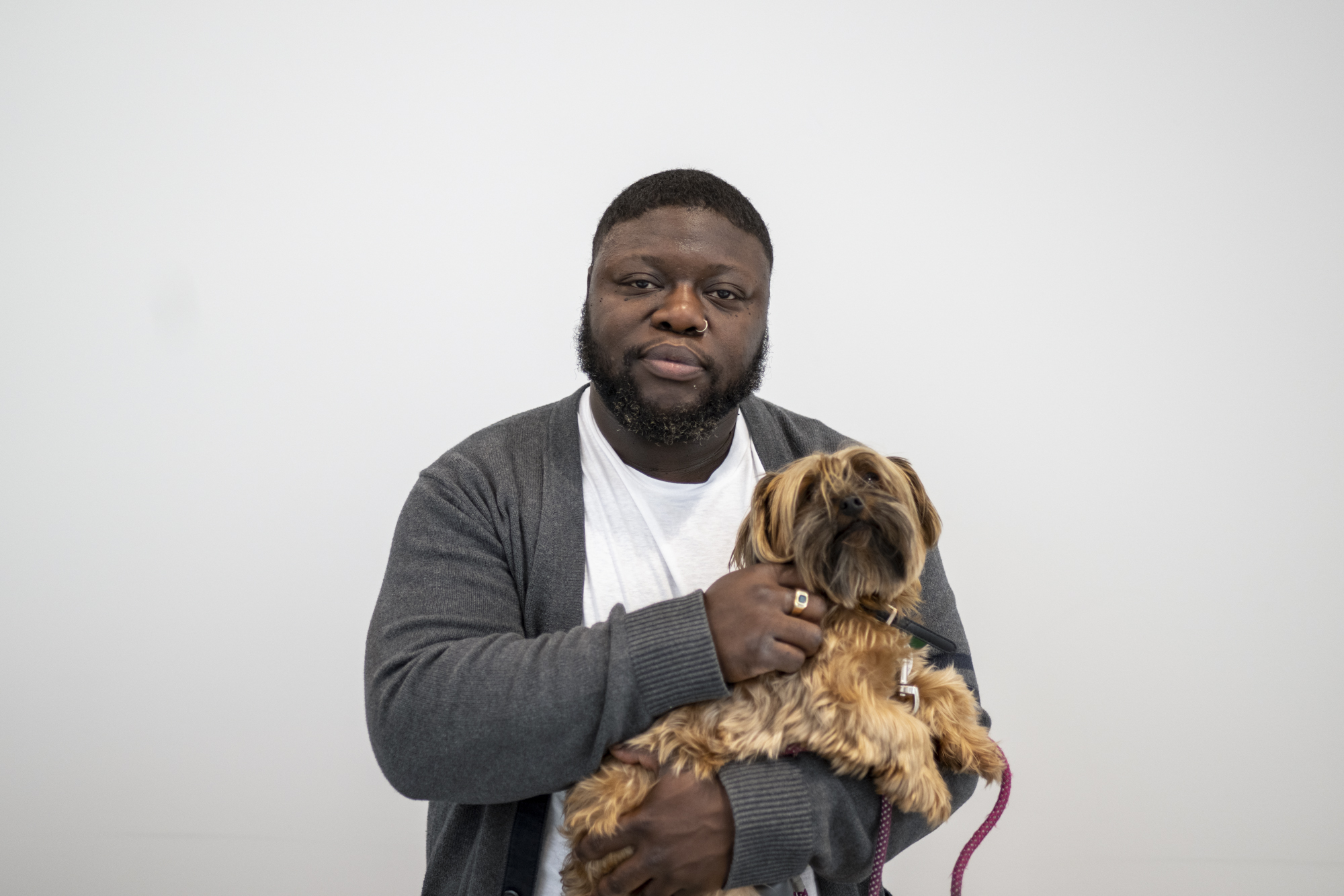A Black man with short black hair and a sort of long black beard and a nose-ring holds a small dog and stands against a blank wall. He wears a white T-shirt and a gray cardigan sweater.