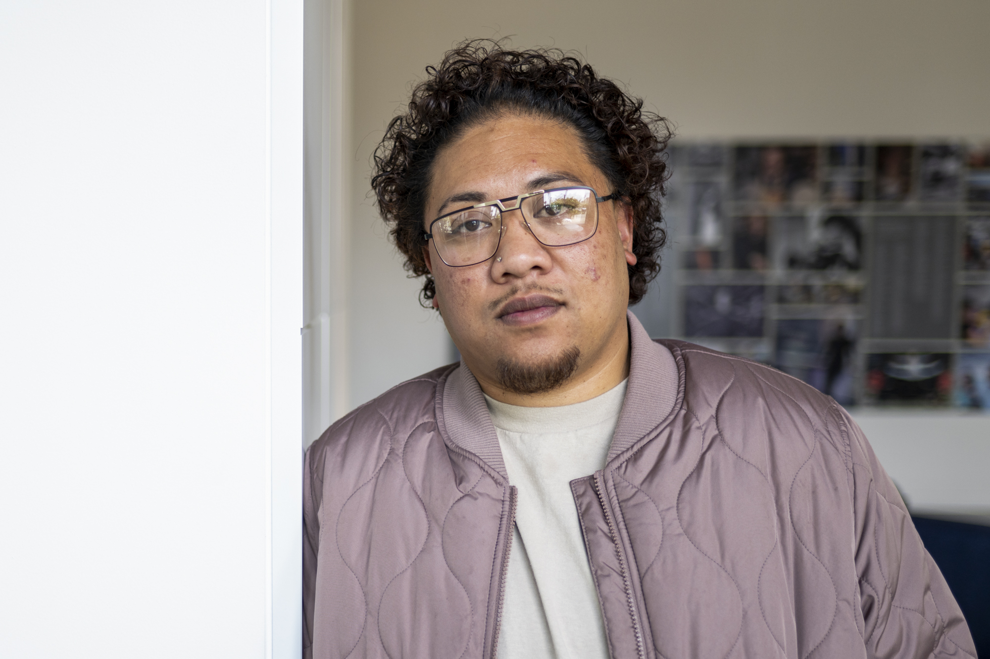 A young Samoan descent man wearing large glasses with a goatee and moustache and curly brown hair leans against a wall in a quilted, lavender bomber jacket and alight gray T-shirt.