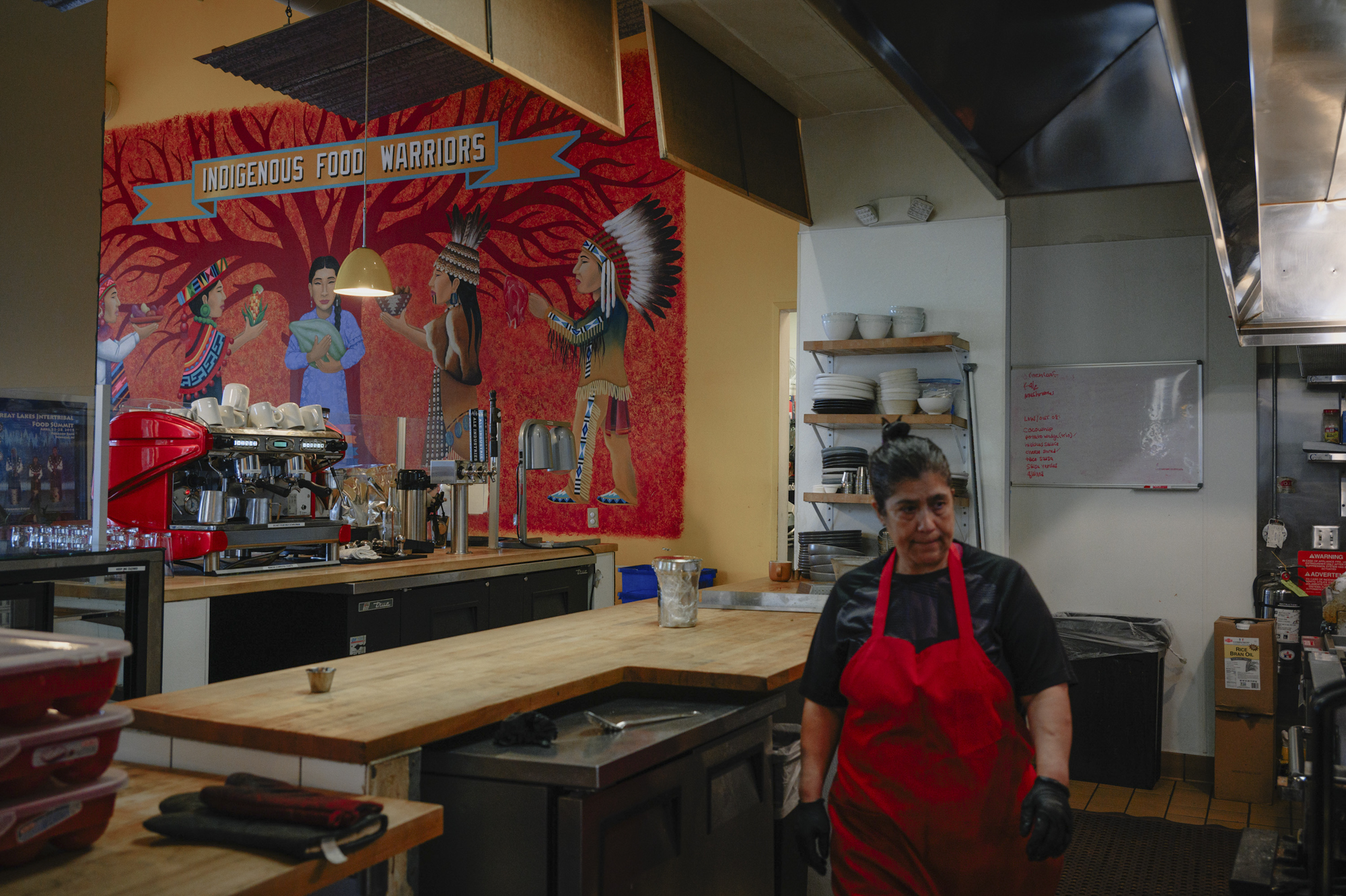 A woman in a red apron walks through a room on one wall of which a mural is painted of people wearing a variety of indigenous clothing and above which the words "INDIGENOUS FOOD WARRIORS" is written.