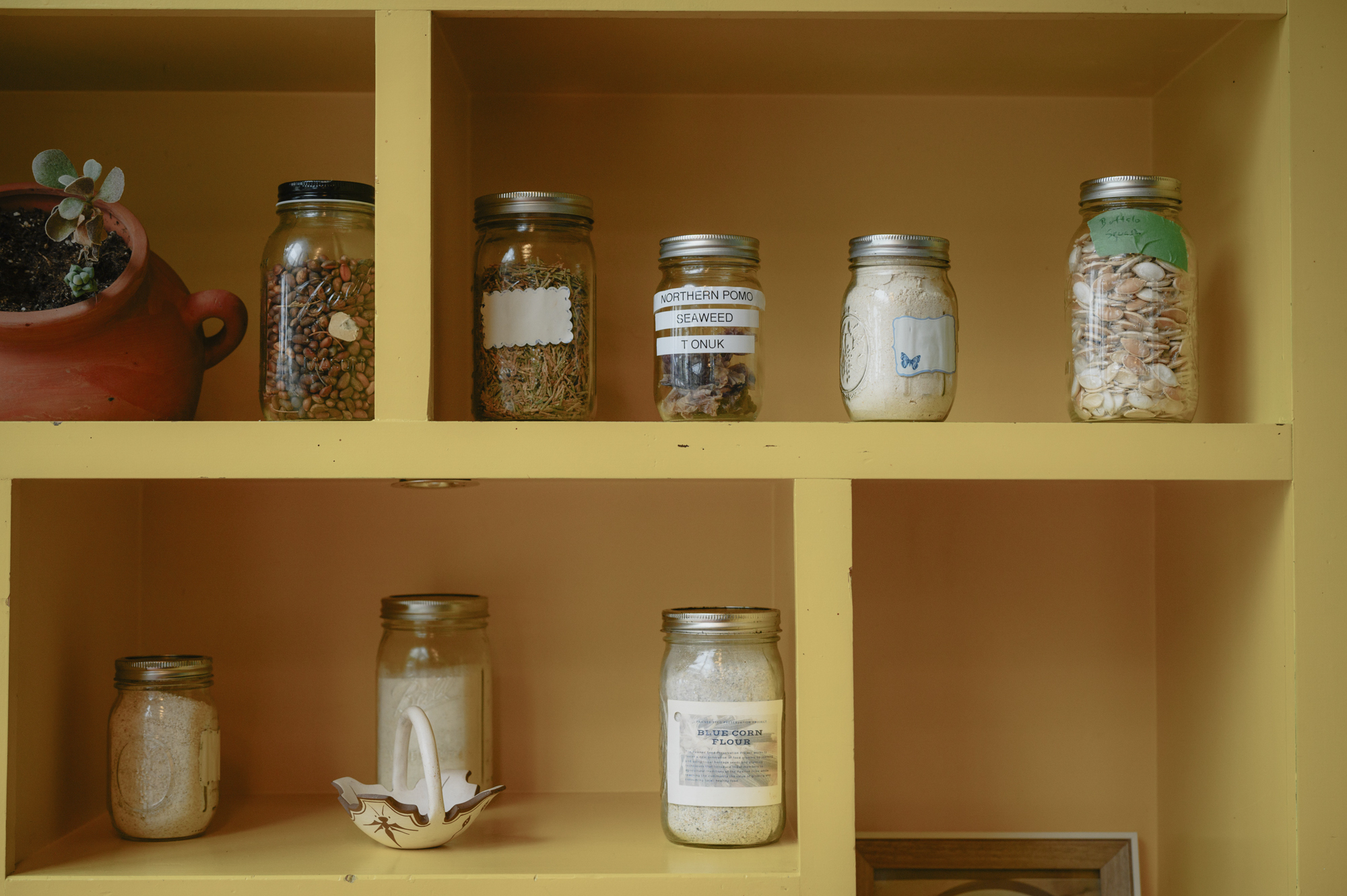 Mason jars filled with various substances -- seeds, powders, herbs -- are aligned on bright yellow shelves