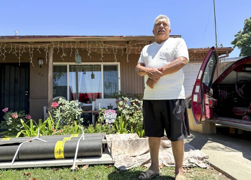 An older Latino man wearing a white t-shirt and black shorts stands in front of a house with a garden, pipe materials and a vehicle on the right.