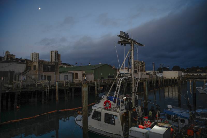 The moon sets as people work on a boat in a harbor in the early morning light.