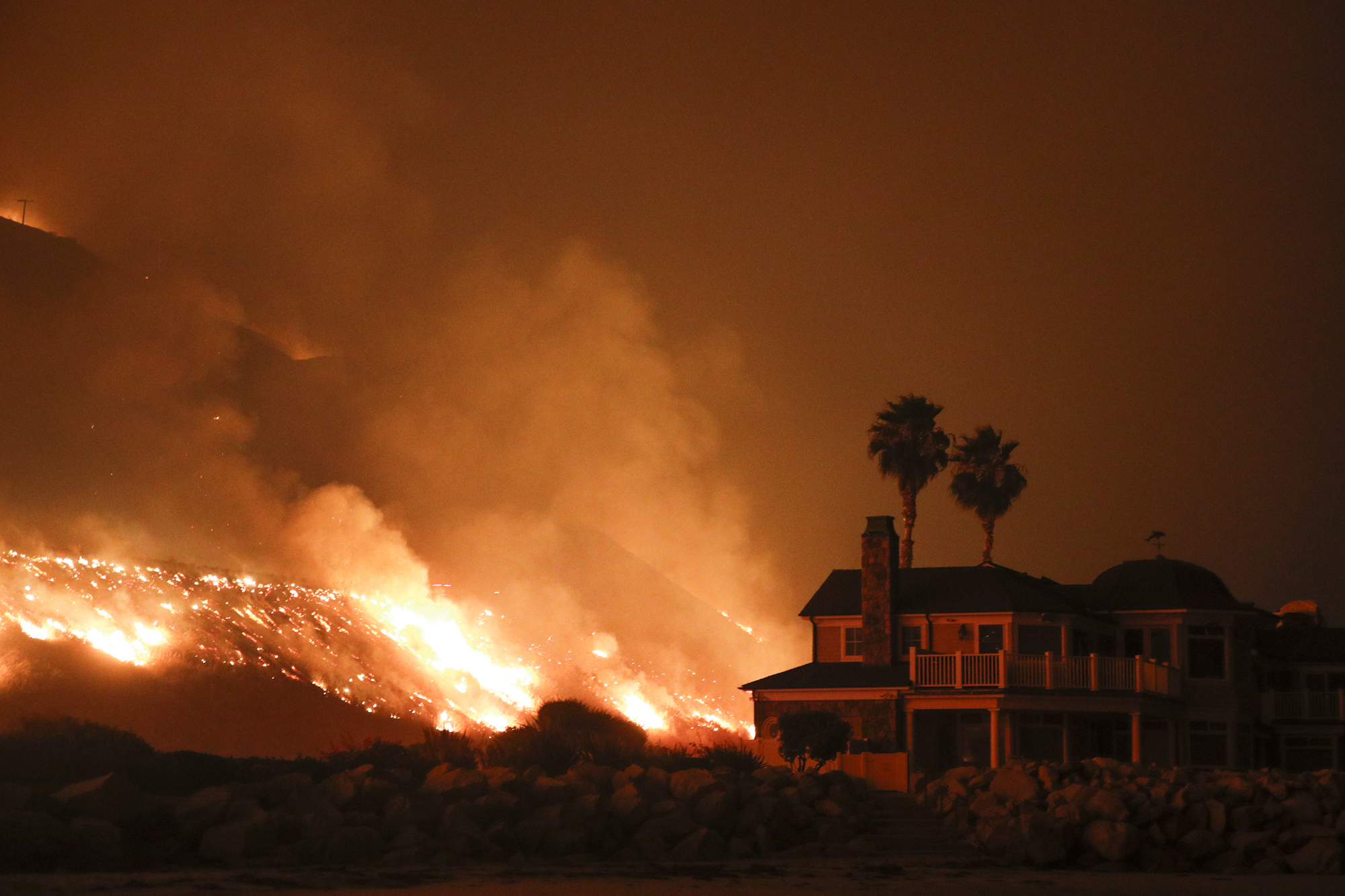 A wildfire rushes toward a mansion in Southern California with two palm trees seen in the home's backyard. The sky is black and the fire glows an ominous, bright orange and red.