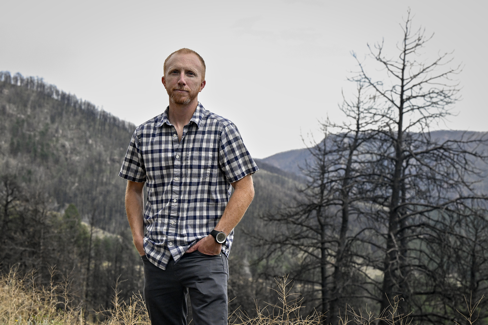 A man with a black and white checkered, button-up shirt, stands amid mountain and trees in Colorado.