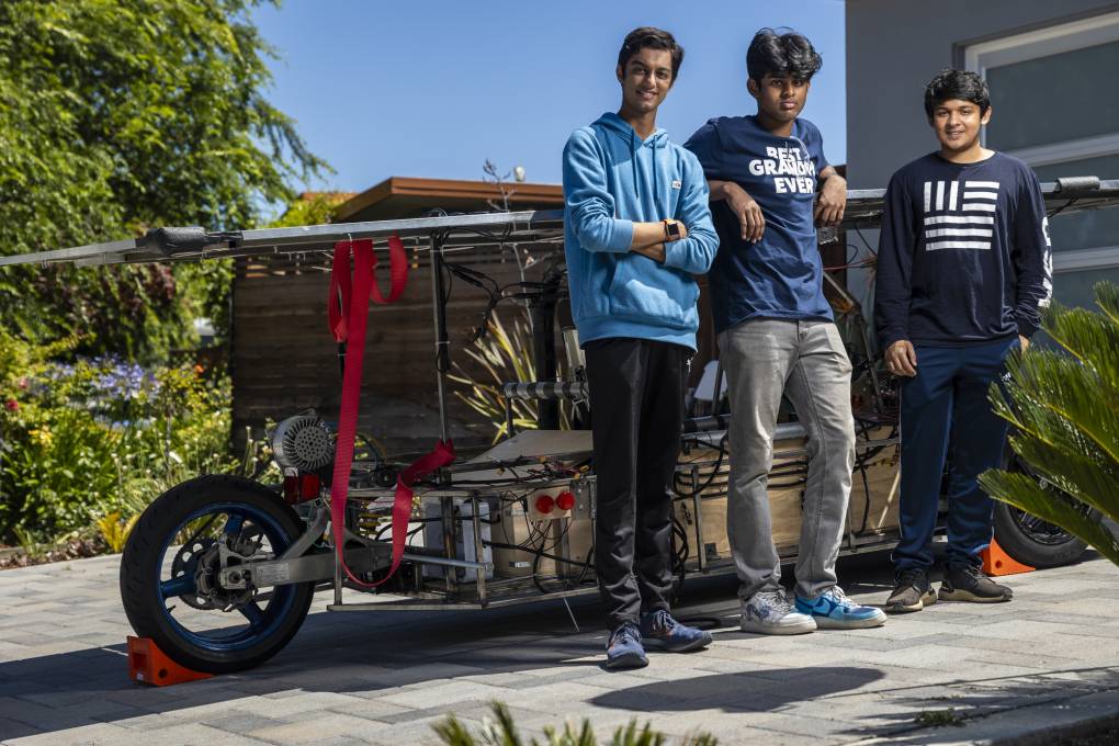 Three high school students lean against a large metallic structure in the driveway of a home.