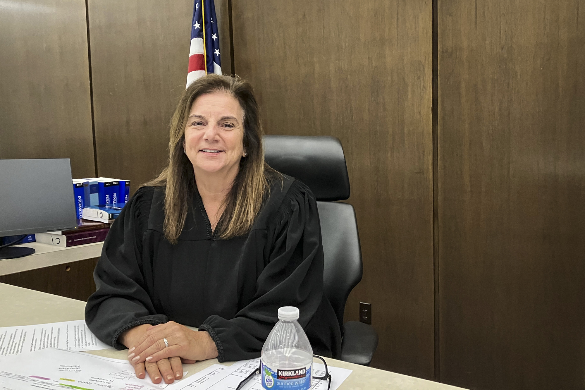 A light-skinned middle-aged woman with long brown hair and wearing black judge's robes smiles at the camera from behind a desk.