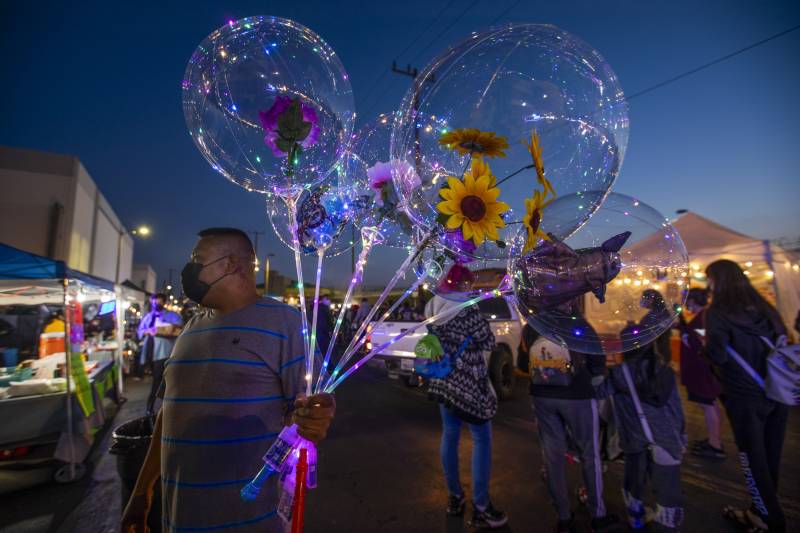 A man holds transparent balloons near outdoor stalls after dark as people pass.