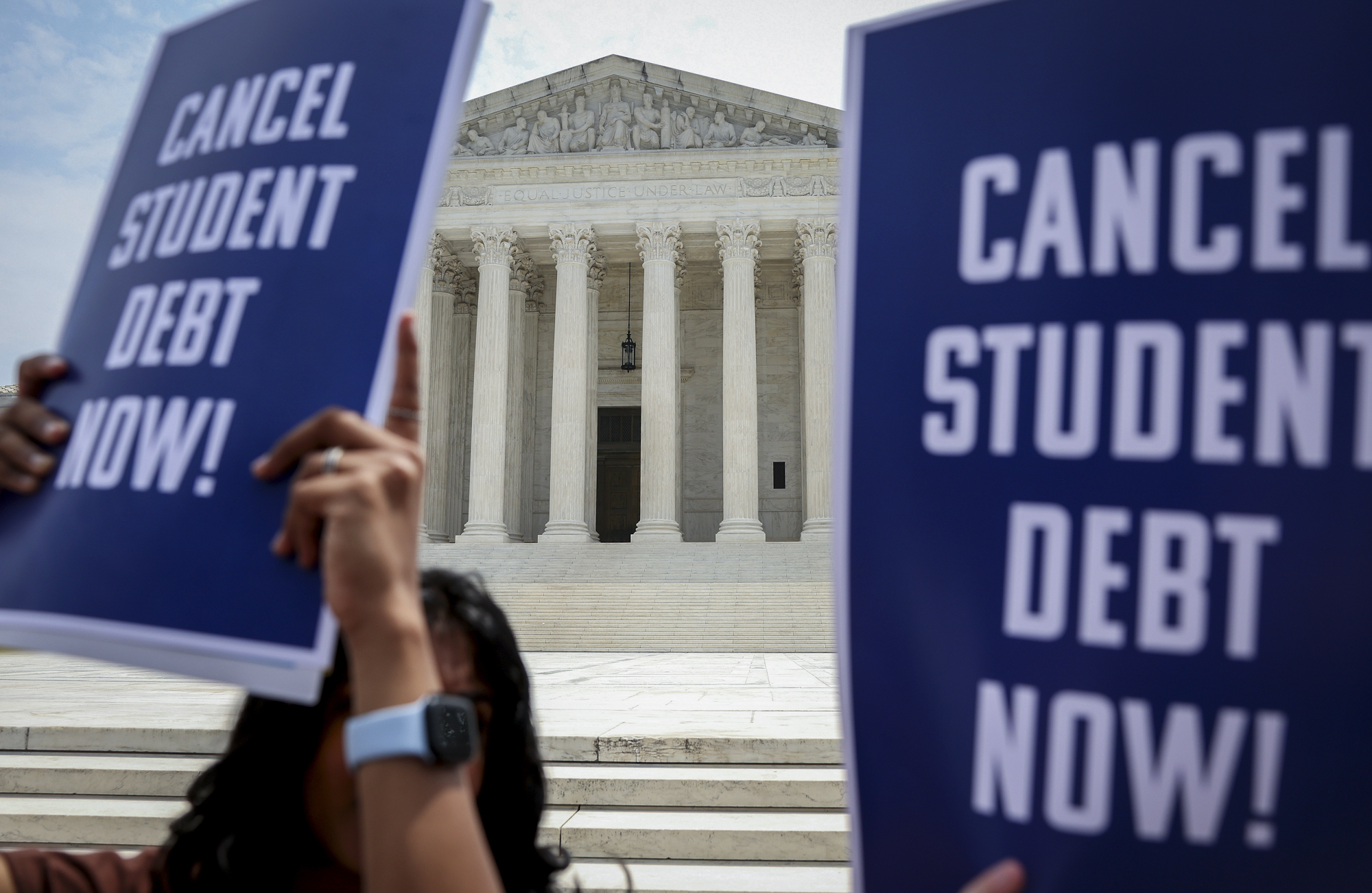 People hold signs reading "Cancel Student Debt Now!" in front of the columned facade of the supreme court.