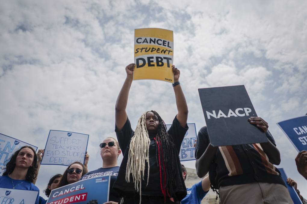 A person with long hair and a black t-shirt holds up a bright yellow sign reading "Cancel Student Debt" amidst others doing similar.