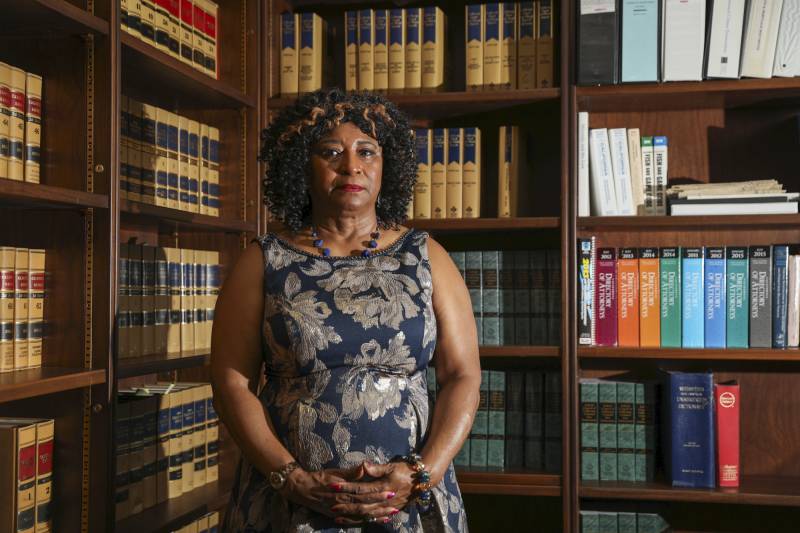 A Black woman wearing a dark, sleeveless floral-printed dress, stands with her hands together in an office filled with books.