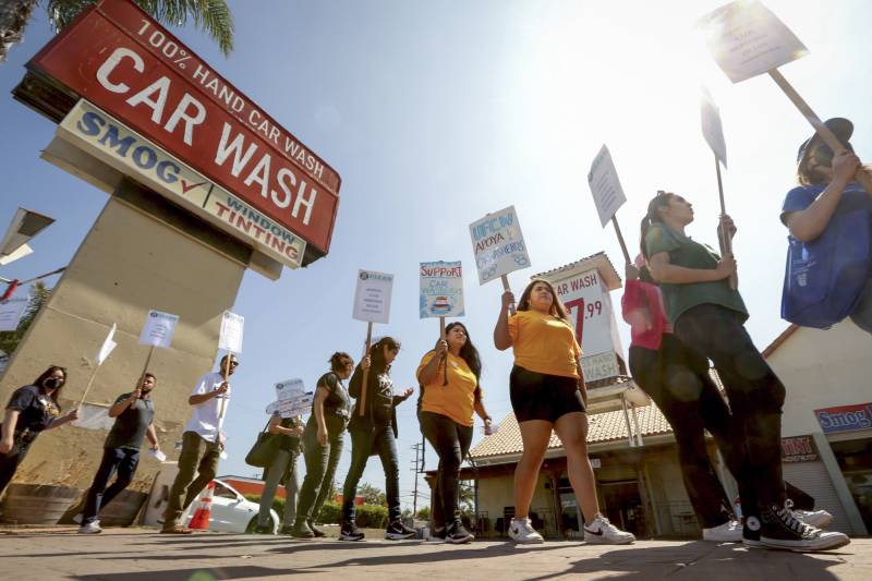 A group of people holds signs and walks in a line in front of a car wash on a sunny day.