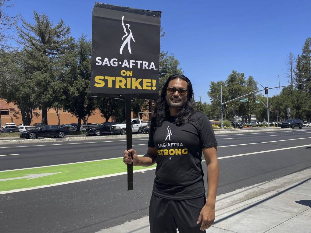 A south Asian man with long hair holds a sign and wears a t-shirt that both read "SAG-AFTRA on Strike"
