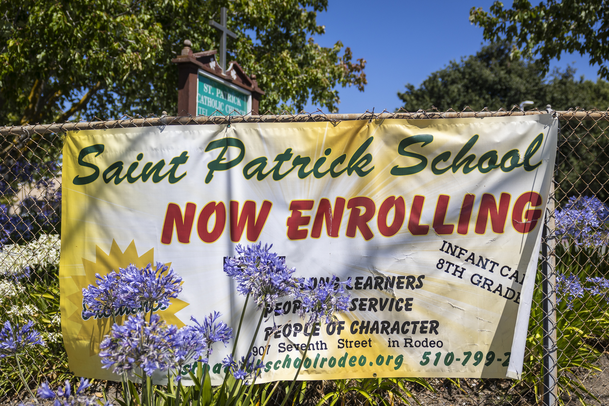 A brightly colored sign hanging on a chain link fence that reads "Saint Patrick School Now Enrolling."
