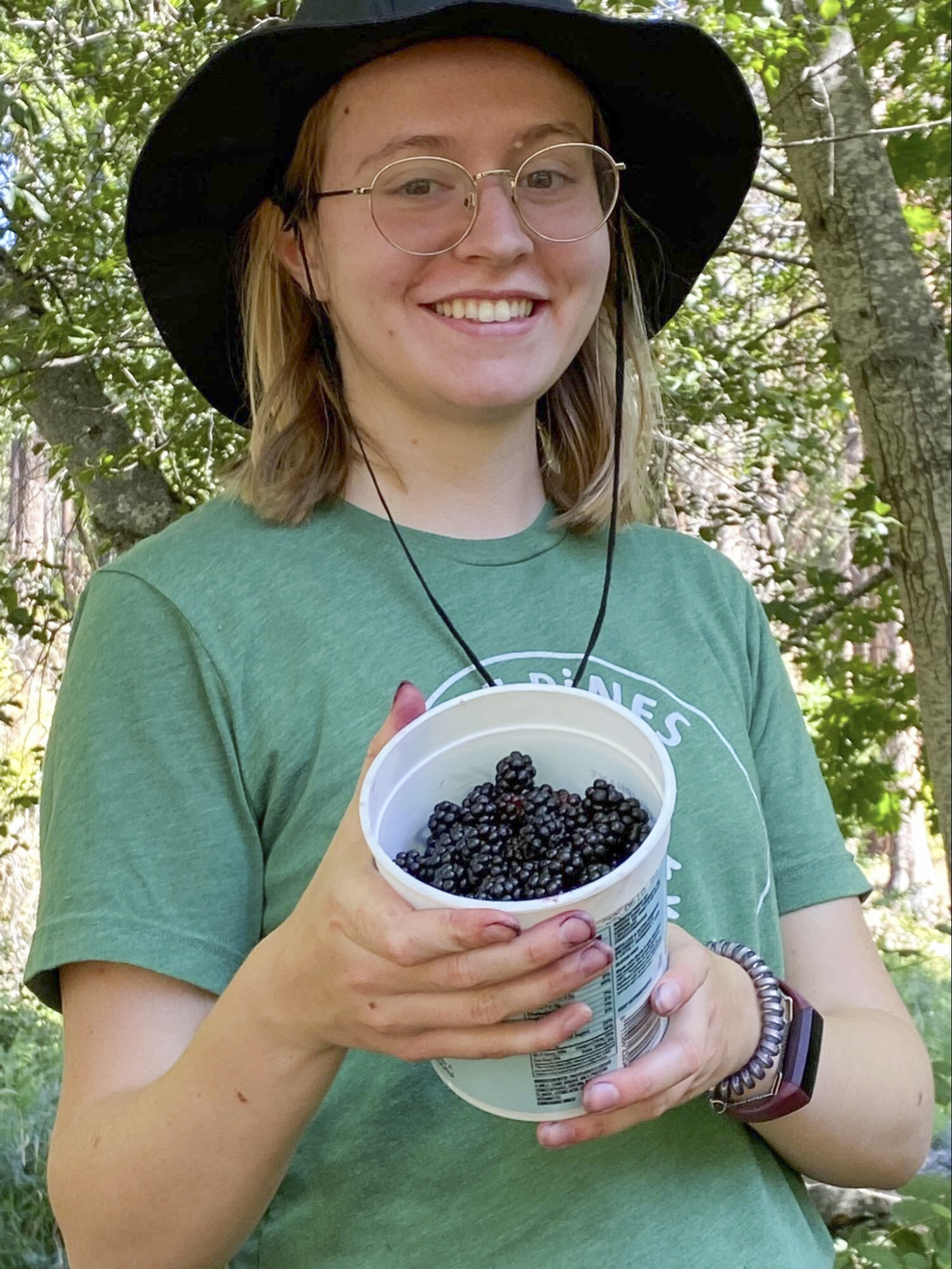 A person with glasses holds a plastic container full of blackberries in a wooded area.