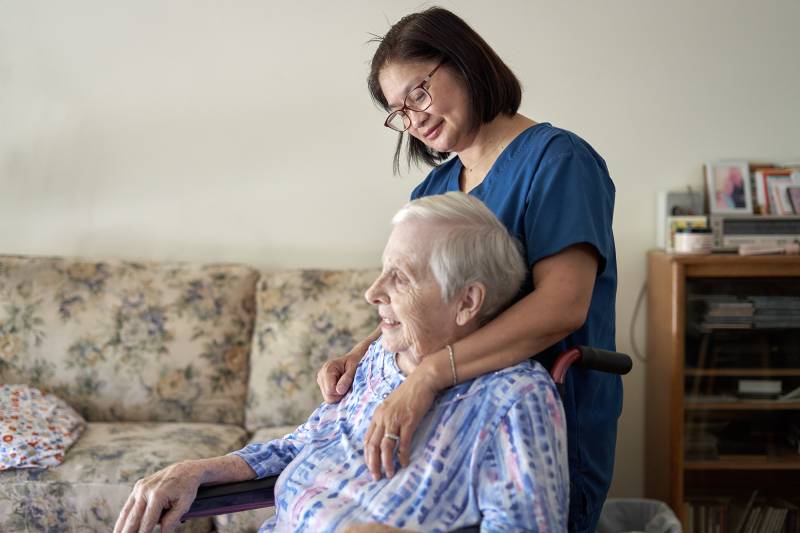 A caregiver in scrubs is affectionately draping her arms over the shoulders of an older woman with gray hair who is sitting in a wheelchair. They're both inside the client's home. A cream-colored, floral-patterned couch is seen in the background.