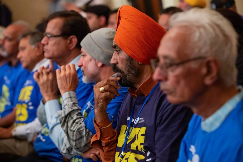 A row of men with tan skin are all wearing blue T-shirts as they sit inside California's Assembly Judiciary Hearing. One man wears an orange headwrap, another a tan beanie. All are seated and looking forward as if listening to a speaker off camera.