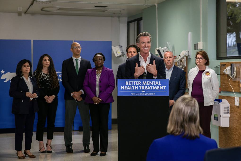 California Gov. Gavin Newsom in a navy blazer and white shirt speaks from behind a black podium with a sign on the front that reads, "Better Mental Health Treatment." Eight men and women in business attire stand solemn-faced behind him.