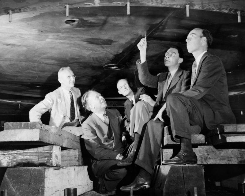 Black and white archival image of 5 men in suits sitting beneath a large, round metal machine, looking up at it.