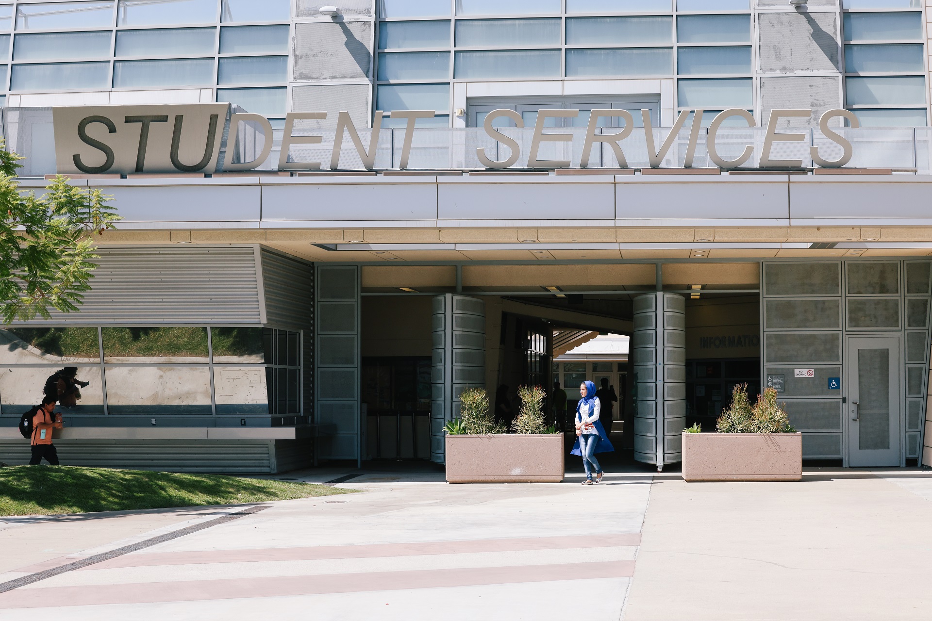 The outside of a community college student services building. It's a large, gray building with many windows. College students are seen with backpacks entering and exiting the building on a sunny day.