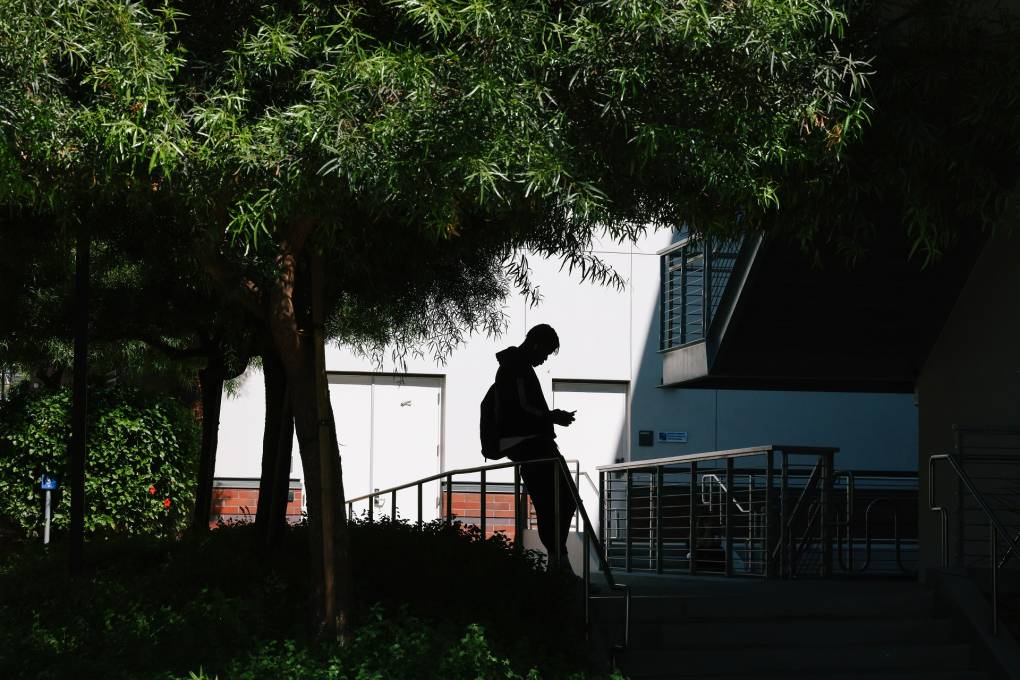 In the shadows under a shady tree, a community college student is seen on their phone. They're wearing a bulky backpack. They're leaning against some staircase rails.