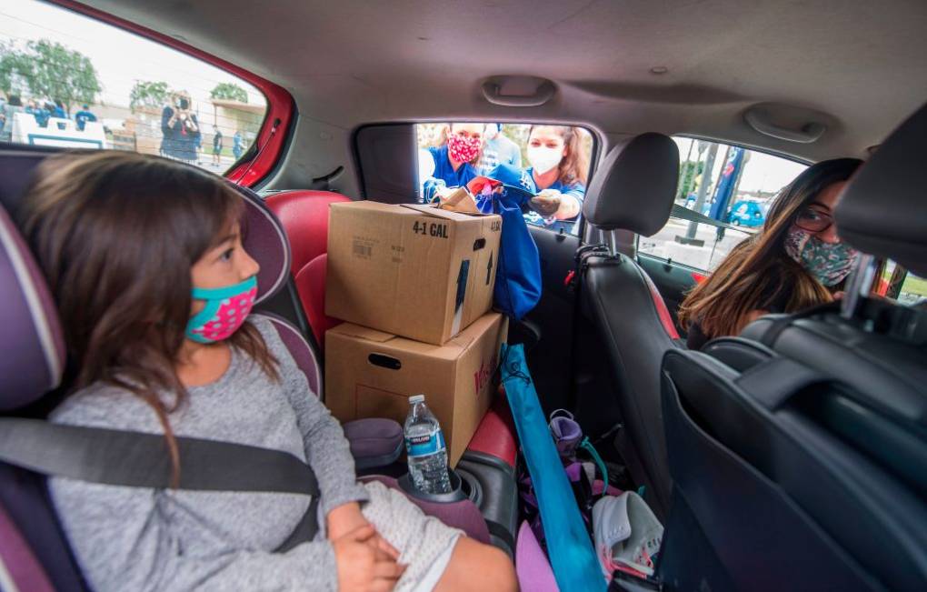 A young girl wearing a pink and turquoise face mask sits in the back of a car next to two cardboard boxes while she looks toward two people wearing masks outside of the car. A woman wearing a mask sits in the drivers seat.