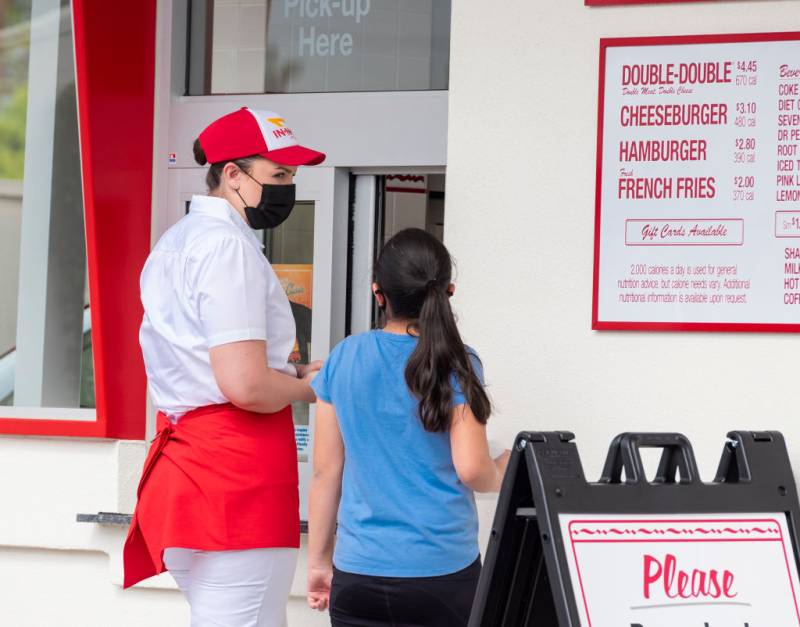 An In-N-Out restaurant worker, wearing a mask and uniform, stands outside the drive-through window taking an order from a customer.