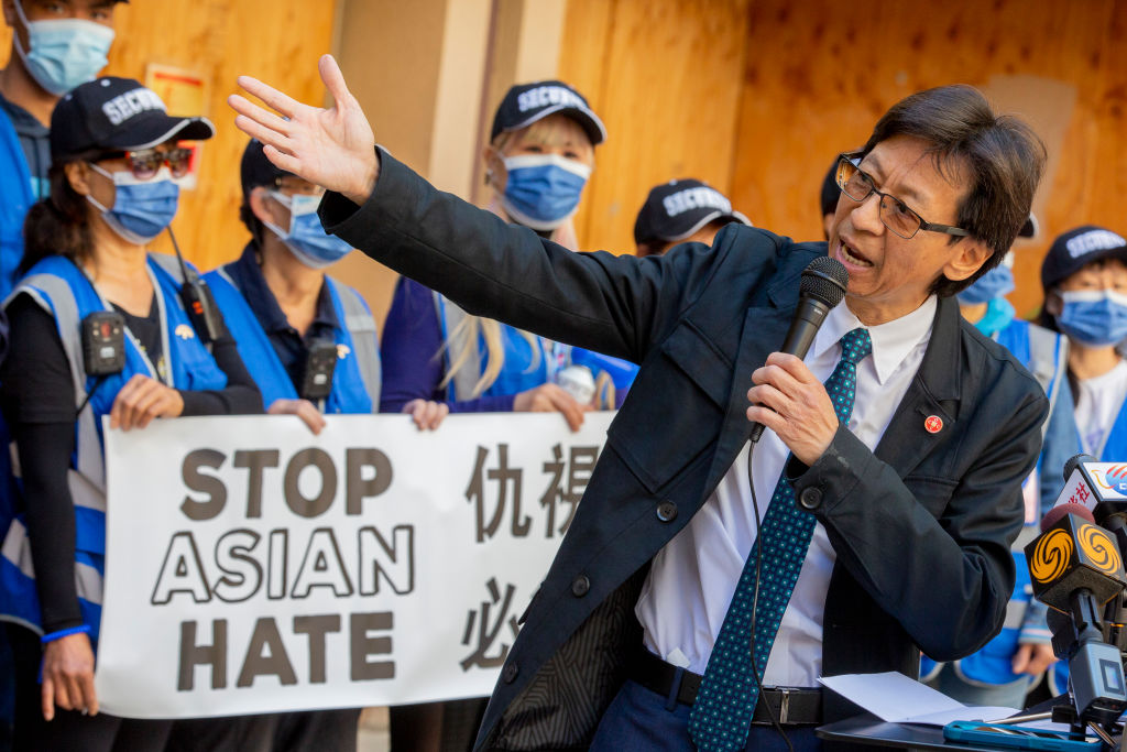A middle-aged Asian man in a suit stands in front of a group of people holding a sign that says 'Stop Asian Hate'